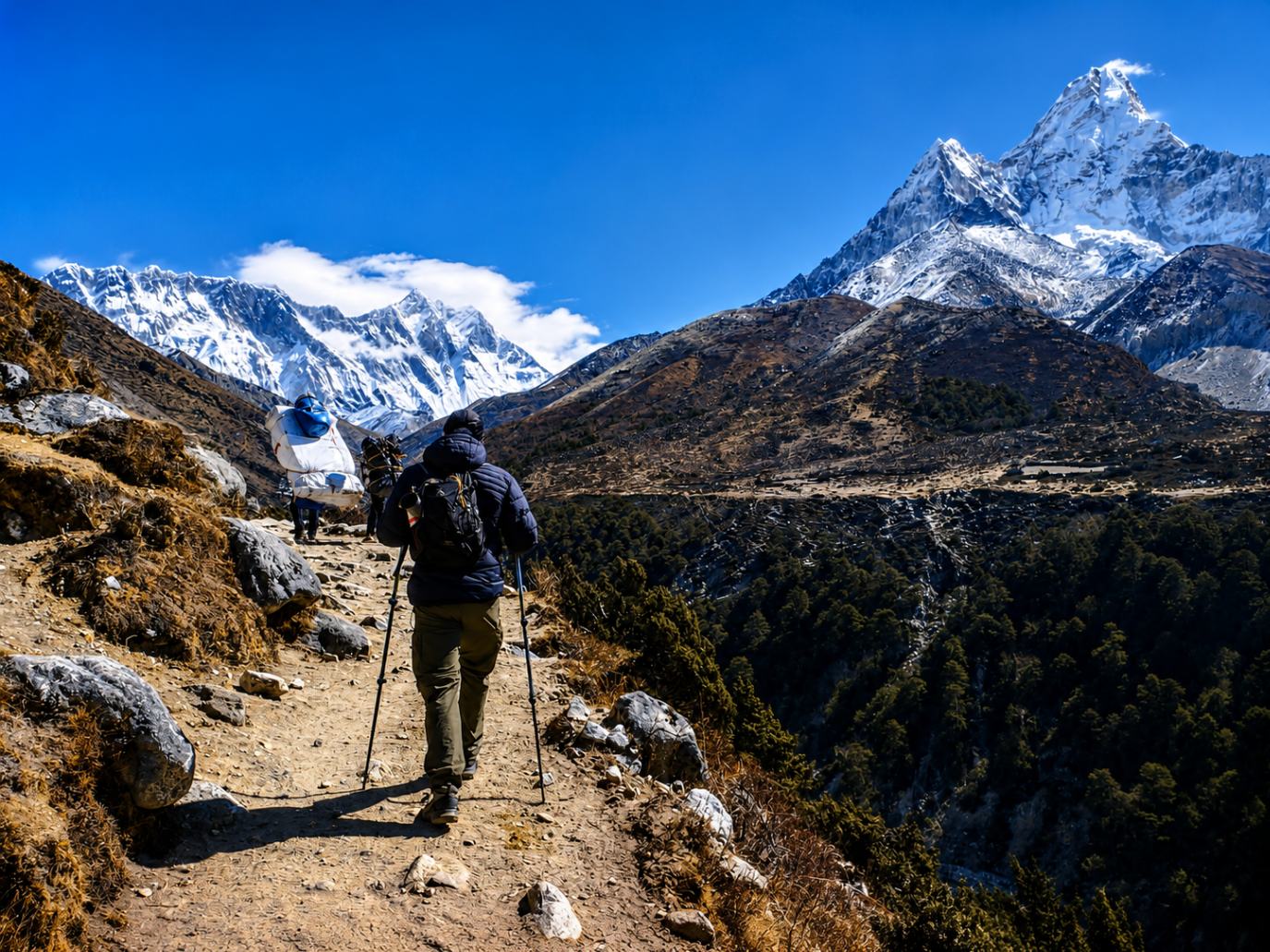 A trekker and a porter walking along the Everest Base Camp trekking trail with panoramic views of snow-capped Himalayan mountains.